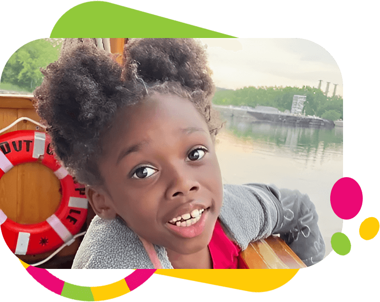 A young child with a wide smile leans over a wooden rail on a boat. A red life preserver is in the background, and water and trees are visible on the other side. The image has colorful graphic elements.