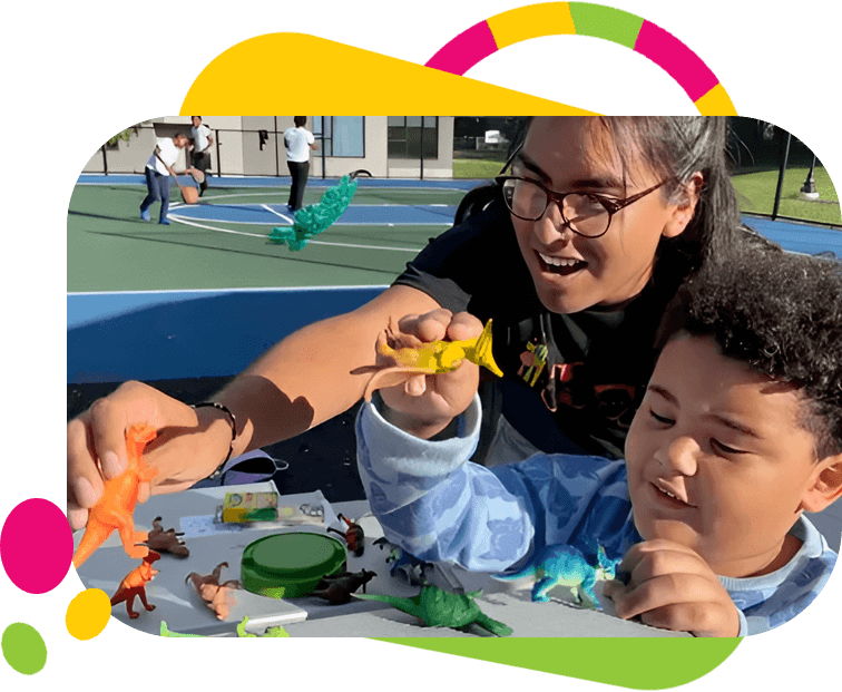 An adult and child enthusiastically play with toy dinosaurs on a table, with others interacting in the background on a sports court.