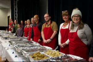 Group of people in red aprons standing behind a table with trays of food, appearing ready to serve at an event.