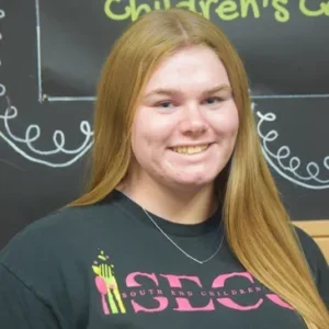 A person with long blonde hair, wearing a black "South End Children's" T-shirt, smiles in front of a chalkboard with some writings and doodles.